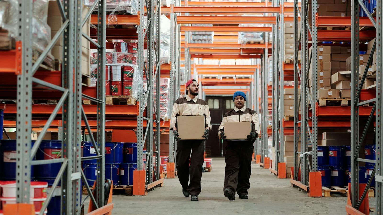 Two workers carrying boxes walk between metal shelves in a spacious warehouse.